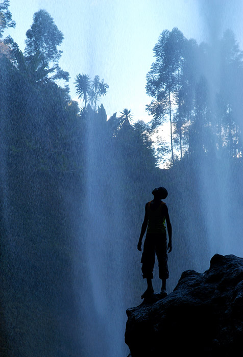Boy standing under Sipi Falls in Uganda Africa by travel photographer Kira Vos (Horvath).